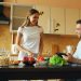 Attractive couple chatting in the kitchen early morning. Handsome man using tablet while his girlfriend cooking breakfast at home