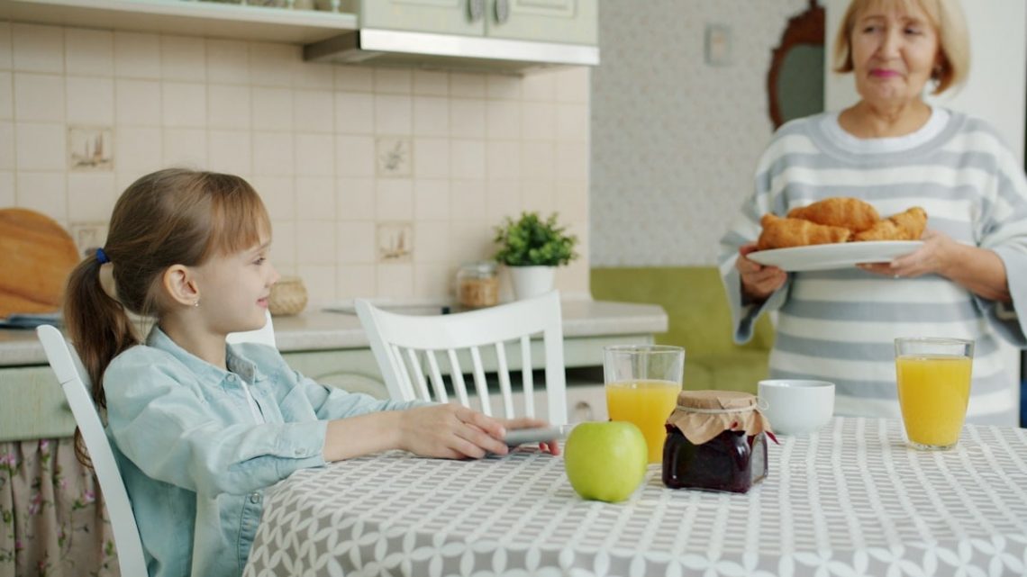 Caring grandmother is bringing plate with fresh croissants feeding granddaughter at home in kitchen, child is clapping hands and smelling pastry