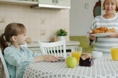Caring grandmother is bringing plate with fresh croissants feeding granddaughter at home in kitchen, child is clapping hands and smelling pastry